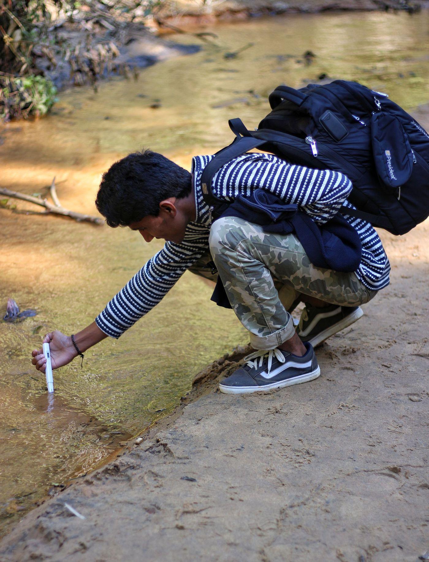 A researcher crouching at the edge of a stream to collect a water sample for field testing